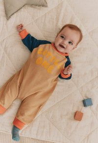 Tan jumpsuit med blå ærmer og orange kant, med store bogstaver "LITTLE ONE." Baby har kort hår og ligger på et struktureret tæppe.