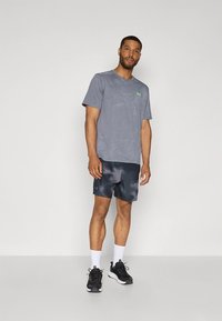 Harmaa urheilupaita vihreällä logolla, yhdistetty tummiin kuviollisiin shortseihin. Mallilla valkoiset sukat ja mustat lenkkarit. Yksinkertainen muotoilu, mukava istuvuus.