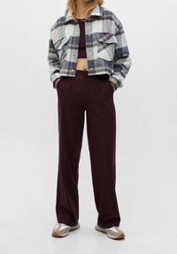 Personne portant une veste écossaise courte sur un crop top bordeaux foncé et un pantalon large bordeaux, assortis à des baskets blanches et beiges.