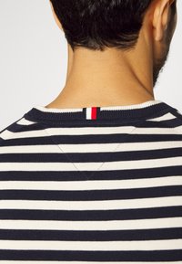 Marineblauw en witte gestreepte trui met ronde hals, met een V-vormige stikseldetail aan de achterkant, voorzien van een klein accentlabel in rood, wit en blauw.