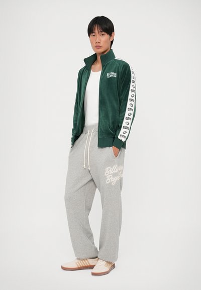 Casaco verde de veludo com fecho e detalhes do logotipo brancos, acompanhado de um par de calças de moletom cinzentas com texto bege. Sapatos bejes com solas castanhas.