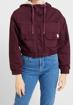 Kvinde iført en kort mørkelilla hættetrøjejakke med frontlommer og blå højtaljede jeans mod en ensfarvet baggrund.