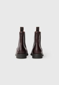 Une paire de bottes Chelsea en cuir marron foncé avec des côtés élastiques noirs et des tirants de marque, montrées de l'arrière sur un fond blanc.