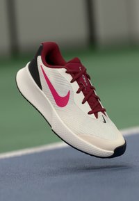 Nike treningssko med en hvit og rød mesh-overdel, rosa swoosh-logo, svarte hælaksenter og teksturert gummisåle, vist i luften.