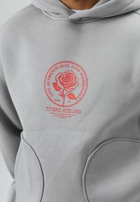 Lichtgrijze hoodie met een centrale rode afbeelding met een roos en tekst, waaronder "YOUNG ATELIER." Zachte stof met een comfortabele pasvorm en grote zakken.