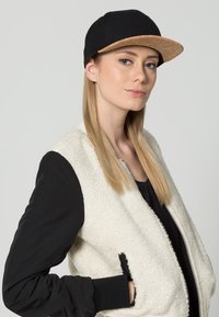 Fekete sapka parafa szélekkel, ami egy krémszínű plüss bomber dzsekihez van párosítva, fekete ujjakkal. A modell semleges arckifejezést mutat, és egyenes haja van.