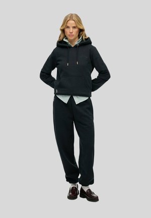 Black hooded sweatshirt with a front pocket, paired with dark sweatpants. Model wears a light striped shirt underneath. Brown shoes.