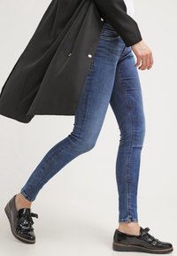 Blå denim skinny jeans med en lätt blekning, ihop med svarta lackskor med en dekorativ rosett. En lång svart kappa hänger över.