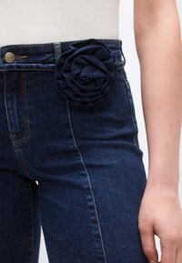 Σκούρο μπλε τζιν υφασμένο με υφή και μεγάλο υφασμάτινο τριαντάφυλλο διακοσμητικό στη μέση. Ορατά κουμπιά στο μπροστινό μέρος.