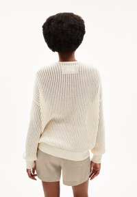 Pull en tricot de couleur crème, à coupe ample, avec des côtes verticales, des manches longues et un col rond. Porté sur un short beige.