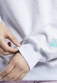 Grå tröja med ribbade muddar, som visar en текстurerad tyg. En liten broderad design i teal syns nära mudden.