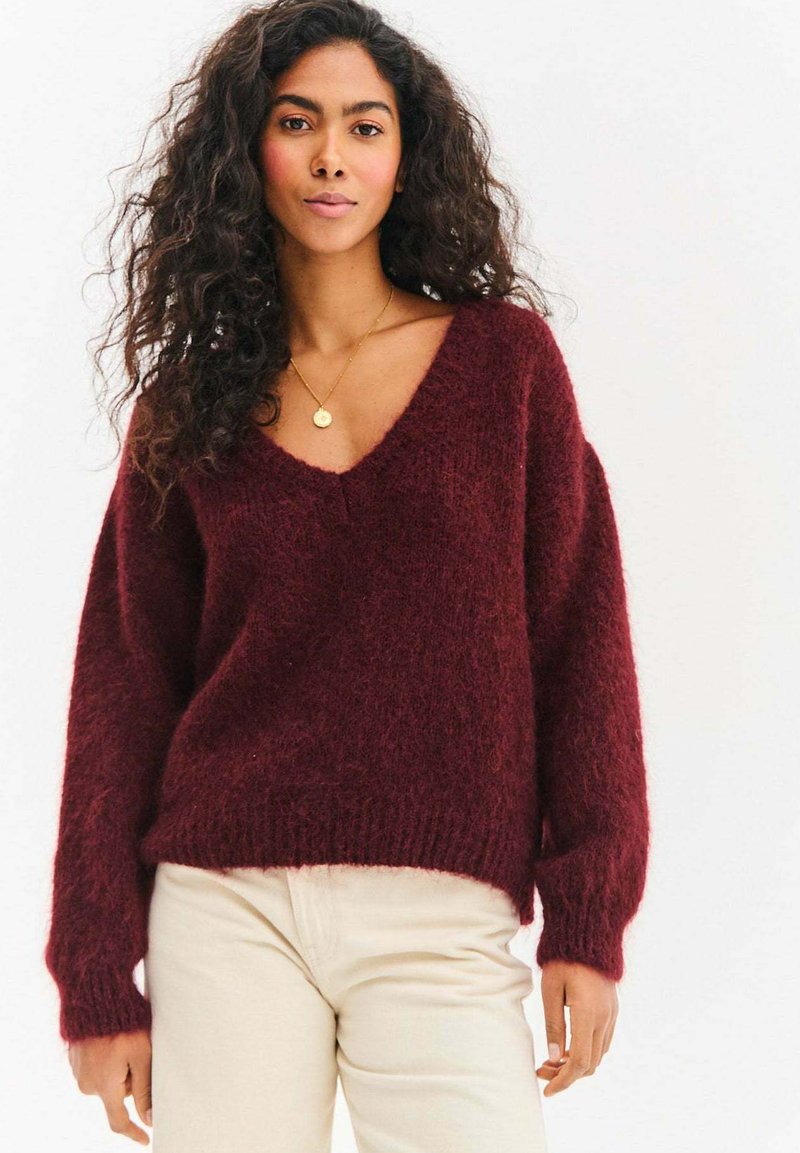 Bordowy dzianinowy sweter z głębokim dekoltem w serek, długimi rękawami i miękką, puszystą fakturą. W połączeniu z kremowymi spodniami.