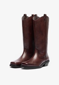Bottines de cowboy en cuir brun avec un bout pointu, une tige haute et des détails de couture subtils. Découpe distinctive haute en haut et un talon bas.