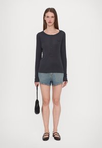 Langärmliges, dunkelgraues Oberteil mit Knopfdetails, kombiniert mit hellblauen Jeans-Shorts. Das Model hält eine schwarze, strukturierte Handtasche.