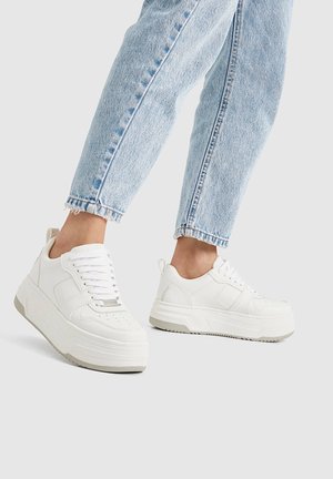 Białe sneakersy na platformie z gładkiej skóry, z teksturowanymi akcentami, sznurowane z przodu, z amortyzującą podeszwą, zestawione z jasnoniebieskimi, skróconymi jeansami.