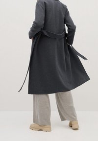 Personne portant un long manteau gris foncé, un pantalon large beige et des bottes beiges, debout avec la ceinture du manteau lâche sur un fond uni.