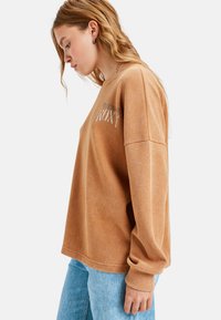 Brązowy sweter o teksturze z luźnym krojem, z srebrnym logo "ROXY" na piersi, w zestawieniu z jasnoniebieskimi dżinsami.