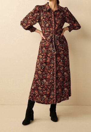 Femme portant une robe midi à manches longues à fleurs avec des épaules bouffantes, une fermeture à boutons sur le devant, une taille nouée, et des bottes à talons noires.