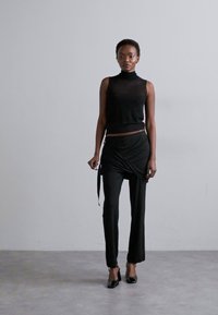 Top preto sem mangas com gola alta, combinado com calças pretas em estilo envolvente. Modelo usa saltos altos pretos, contra um fundo neutro.