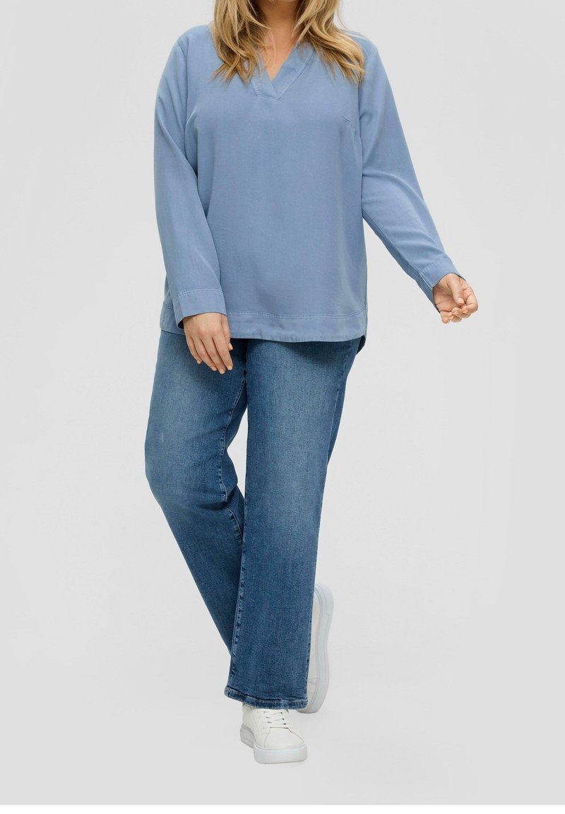 Top bleu clair à manches longues et col en V associé à un jean en denim bleu moyen à larges jambes et des baskets blanches. Tissu lisse, coupe décontractée.