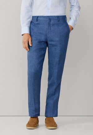 Homme portant un pantalon bleu ajusté, une chemise à rayures bleu clair, et des chaussures en daim marron, debout avec une main dans la poche devant un fond uni.