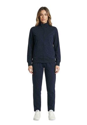 Une femme porte une veste bleu marine zippée et un pantalon assorti, avec des baskets blanches, sur un fond blanc uni.