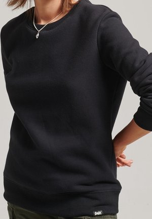 Frau trägt ein schlichtes schwarzes Sweatshirt mit langen Ärmeln und einem kleinen Logotag nahe dem Saum, kombiniert mit olivgrünen Hosen.