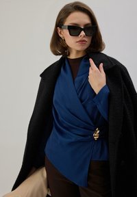 Blusa azul marino drapeada con un diseño retorcido y broche de hoja dorada, superpuesta bajo un abrigo negro. La modelo lleva gafas de sol negras y pendientes dorados.