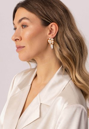 Frau mit welligem hellbraunem Haar, die ein cremefarbenes Satinblazer und goldene Blumenohrhänger mit Perlakzenten trägt, blickt nach links vor einem neutralen Hintergrund.