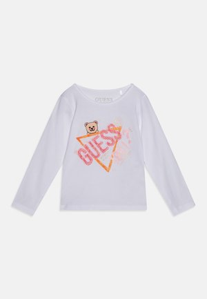 Chemise blanche à manches longues avec un ours en peluche souriant qui dépasse d'un triangle orange et le mot "GUESS" en lettres pailletées roses sur le devant.
