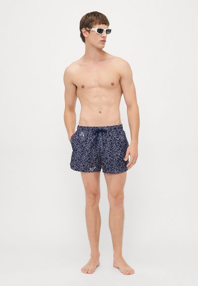 Oberkörperfreier Mann trägt weiße Sonnenbrille und marineblaue Badeshorts mit weißem Fischmuster, steht barfuß vor weißem Hintergrund.