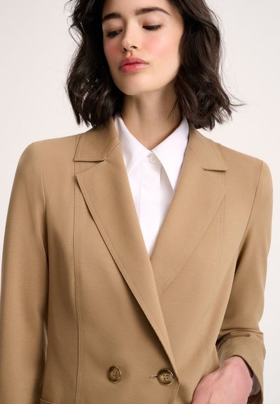Light brown double-breasted blazer with sharp lapels and a smooth texture, paired with a white collared shirt underneath.