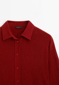 Chemise rouge texturée à manches longues avec col classique et quatre boutons visibles sur fond blanc.