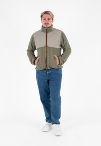 Giacca in pile verde con parte superiore imbottita beige, chiusura con zip e due tasche laterali. Indossata con jeans blu e sneaker bianche.