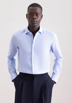 Uomo con camicia da uomo a quadretti azzurri chiari infilata in pantaloni a pieghe blu scuro, in piedi con le mani in tasca davanti a uno sfondo semplice.