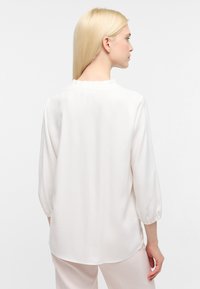 Cremefarbige Bluse mit Dreiviertelärmeln, rundem Ausschnitt mit Rüschendetail und einem glatten, leichten Stoff.