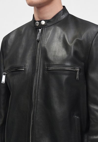 Homme portant une veste en cuir noire avec fermeture éclair devant et deux poches poitrine horizontales zippées, col montant avec boutons-pression.