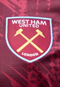 Λογότυπο της ποδοσφαιρικής ομάδας West Ham United με σταυρωμένα χρυσά σφυριά και κείμενο σε φόντο από ύφασμα χρώματος βυσσινί με υφή.