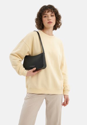 Jonge vrouw met krullend haar, gekleed in een beige sweatshirt en broek, met een zwarte schoudertas met merklogo.