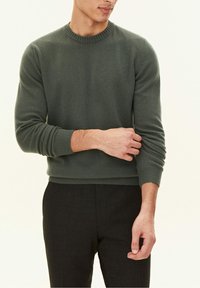 Maglione verde lavorato a maglia con scollo e polsini a coste, indossato da una persona con pantaloni scuri, le mani posate delicatamente sul braccio.