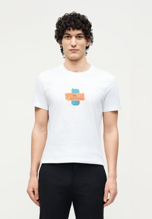 Ung mand med krøllet hår iført en hvid T-shirt med et farverigt abstrakt logo og sorte bukser, står mod en ensartet baggrund.