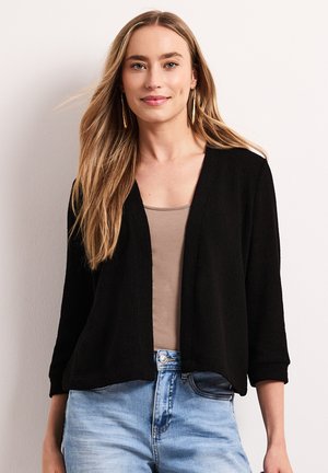 Kobieta z długimi blond włosami, nosząca złote kolczyki koła, czarny otwarty kardigan, beżowy top i jasnoniebieskie dżinsy na jednolitym tle.