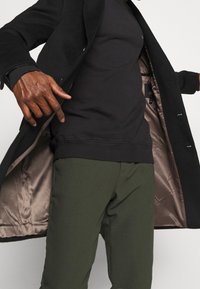 Hombre con abrigo negro abierto que muestra el forro brillante beige, sudadera negra y pantalones verde oliva contra un fondo blanco.