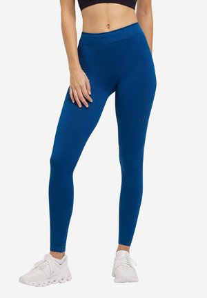 Blaue Leggings aus dehnbarem Material, mit einem hohen Bund und einer glatten Textur. Kombiniert mit weißen Sportschuhen.