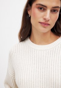 Strickpullover in Off-White mit Rippstrick-Textur, runder Ausschnitt und lässiger Passform. Es werden silberne Creolen getragen.