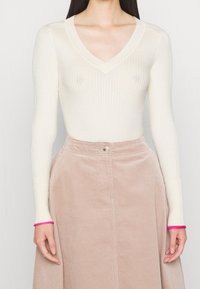 Vrouw in een crèmekleurige geribbelde V-hals trui met lange mouwen die bij de manchetten zijn opgerold, en een high-waisted beige corduroy rok.