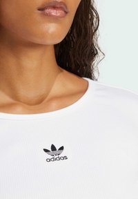 White ribbed shirt with a rounded neckline and a small black embroidered Adidas logo on the left chest area. Smooth fabric texture.
