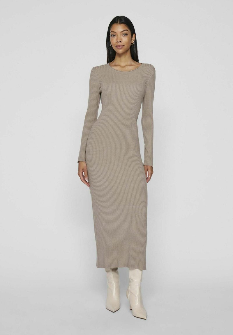 Long-sleeve ribbed dress in light taupe, fitted design, round neckline, reaching mid-calf, paired with cream-colored high-heeled boots.