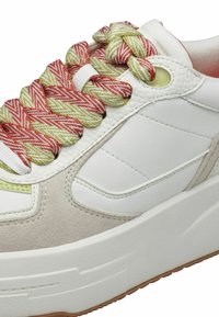 Baskets blanches et beiges avec des lacets torsadés épais rouges et verts passés à travers des œillets dorés sur une semelle en caoutchouc blanche.