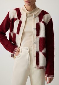 Czerwony i kremowy kardigan w krateczkę o fakturowanym splocie, z okrągłymi srebrnymi guzikami i paskowanymi mankietami, zestawiony z kremowymi spodniami.